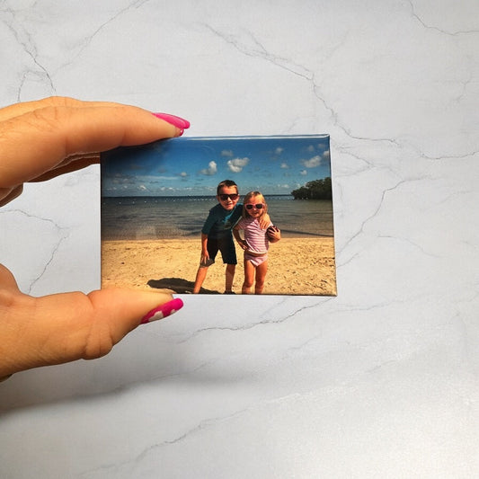Hand holding a custom photo magnet featuring two young children smiling on a sunny beach, wearing sunglasses, with the ocean and blue sky in the background.